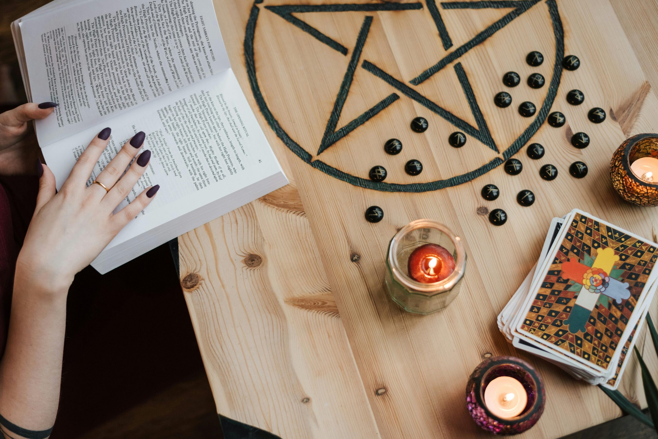 A woman holding a text book, as she sits by her wooden desk. She is conducting a spell with candles, a pentagram and taro cards.