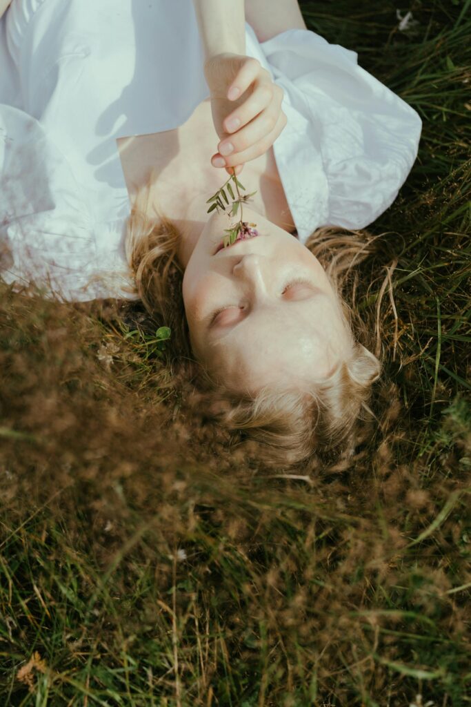 A woman in a white dress laying in an open field, smelling a sprig of flower. The sun is bright and the glow of the suns rays lighten her hair.