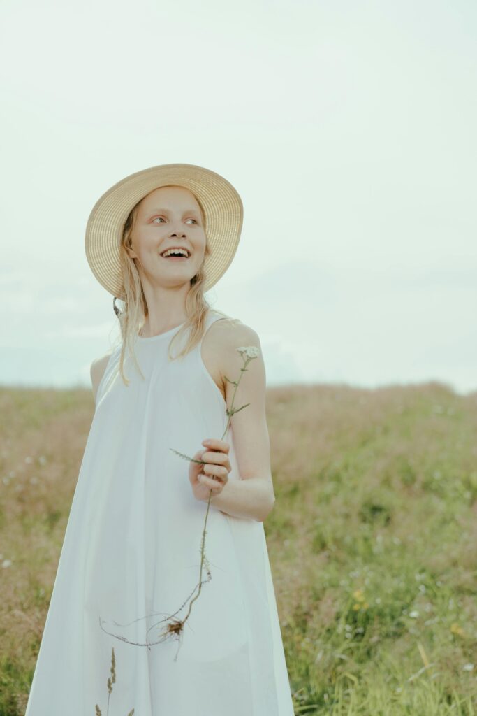 A pale women wearing a white sundress and straw hat smiles brightly while holding a daffodil. She is standing in an open field, the flowers and grass tall and swaying in the wind.