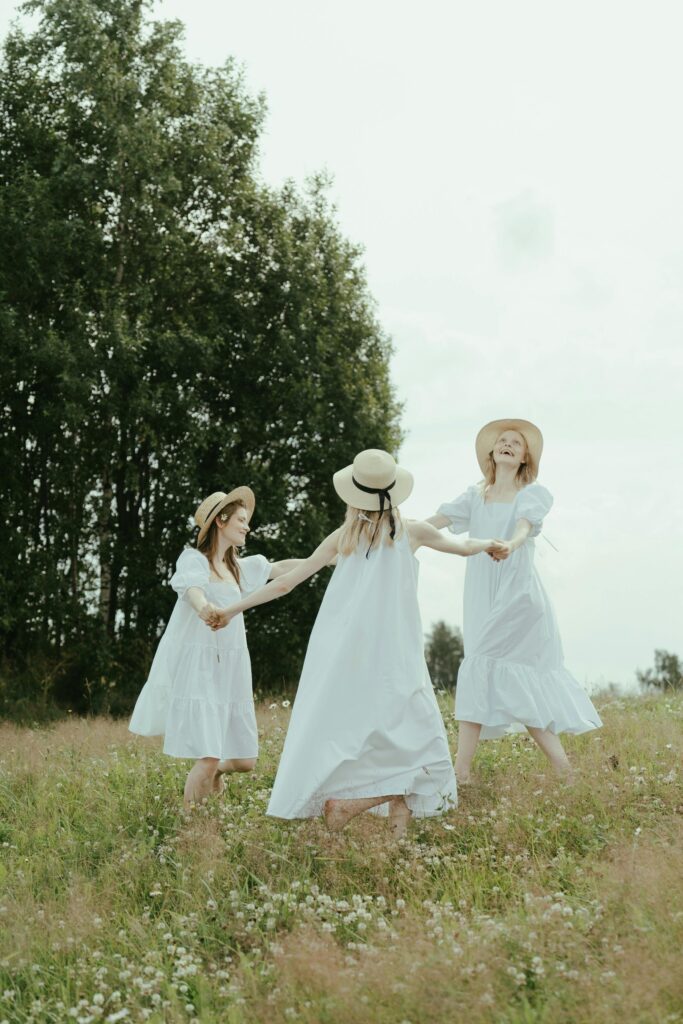 A group of women in a white dresses spinning around in an open field. The sun is bright and the glow of the suns rays lighten their hair.