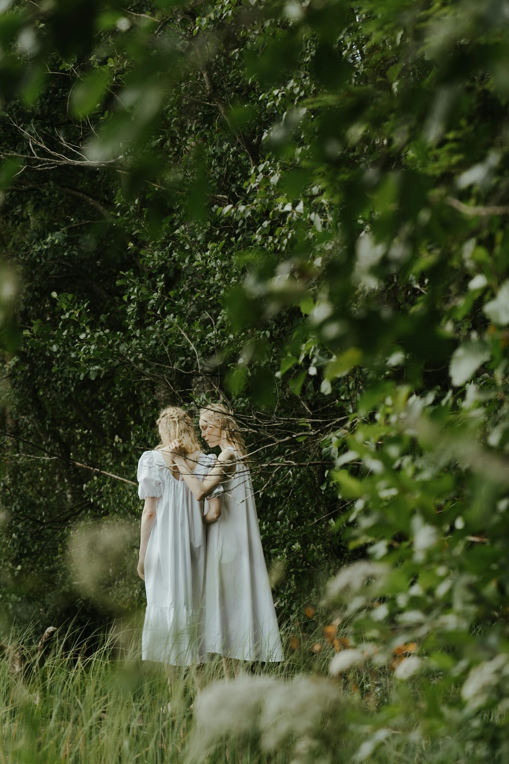 Two pale women in white dresses stand together in a lush green forest.